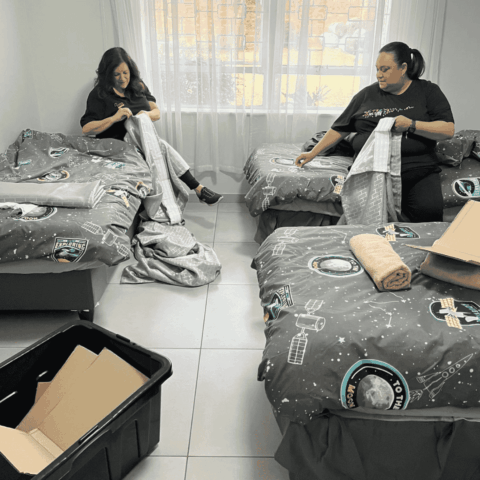 Two women sit on separate beds in a bright room, placing matching space-themed bedding on the beds. A box and a towel are on one bed; an open plastic bin is on the floor in front of them.