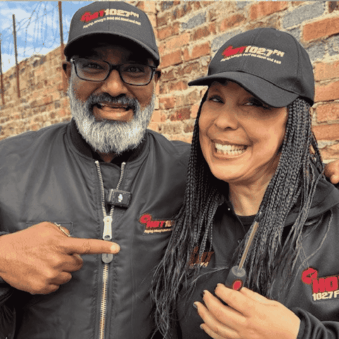 Two smiling people in black Hot 102.7 FM jackets and caps pose together in front of a brick wall. One person points at the other, both wearing glasses and appearing cheerful.