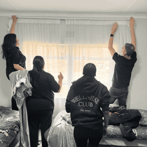 Four people are adjusting white curtains on a window in a bedroom. Two stand on beds to reach the curtain rod, while two others, one wearing a WELLNESS CLUB hoodie, assist from the floor.
