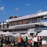 A busy scene outside Wonderboom National Airport shows people in safety vests, tents, and booths under a sunny sky. The airport building has a sign reading WONDERBOOM NATIONAL AIRPORT above the arrivals area.