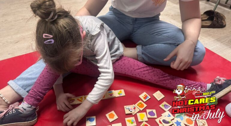 A young girl sits on a red mat playing with colorful wooden tiles while an adult sits beside her. The image has a HOT 102.7 FM Hot Cares Christmas in July logo in the bottom right corner.