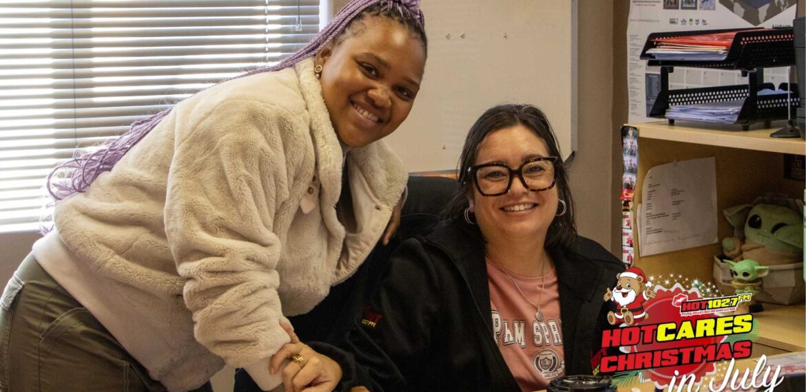 Two women smiling in an office, one standing and one sitting at a desk. A festive HOT CARES Christmas in July logo is displayed in the bottom right corner.