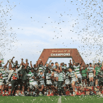 A rugby team celebrates winning a championship on a field, standing under a large CHAMPIONS sign while confetti falls around them. Players and coaches raise their hands in victory and joy.