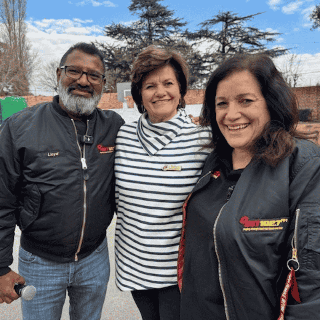 Three adults smile outdoors under a blue sky. Two wear black jackets with yellow and red logos, and the person in the middle wears a white and black striped top. Trees and a brick wall are in the background.