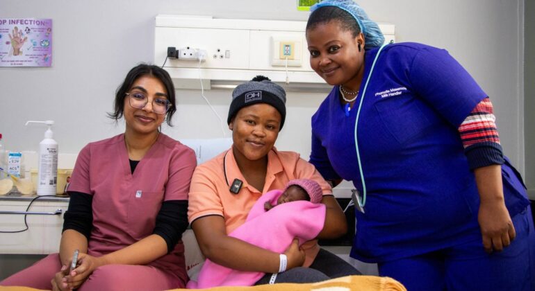 Three women smile at the camera in a hospital room. One woman, wearing a hat, holds a newborn baby wrapped in a pink blanket, while the other two women in medical scrubs offer support and guidance on breastfeeding.
