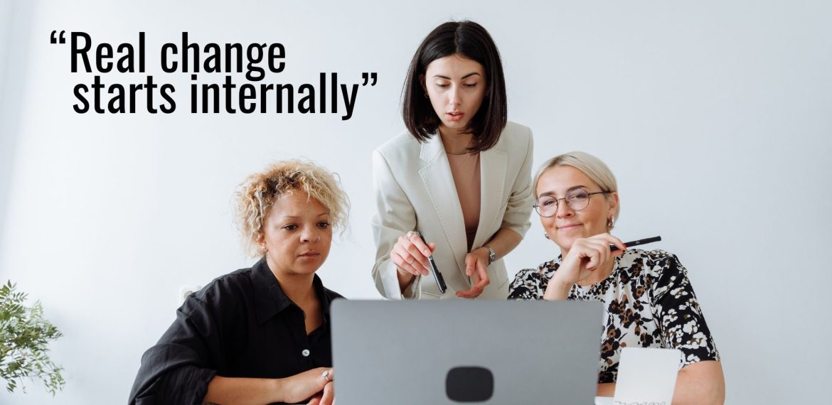 Three women sit together at a desk, focused on a laptop, while one woman stands and points at the screen. The text reads, “Real change starts internally”—a reminder that true progress goes beyond pinkwashing.