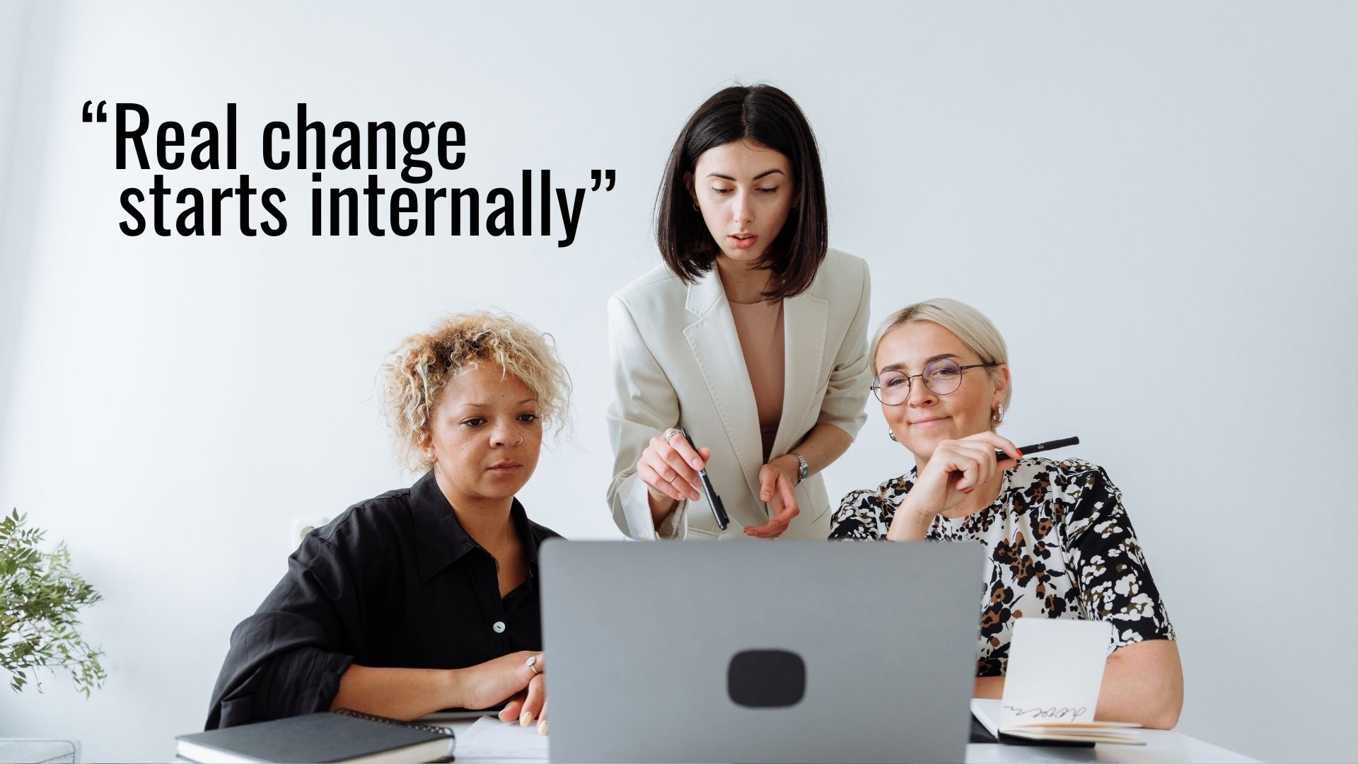 Three women sit together at a desk, focused on a laptop, while one woman stands and points at the screen. The text reads, “Real change starts internally”—a reminder that true progress goes beyond pinkwashing.