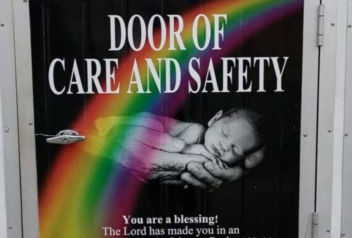 A sign on a door reads Door of Care and Safety above an image of adult hands holding a baby, with a rainbow across the picture. Text below says, You are a blessing! The Lord has made you in an amazing way.