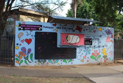 A colorful mural with hearts, flowers, and birds on a wall labeled NEW BEGINNING BABY WALL. A hatch in the wall is shown, surrounded by contact details and caring messages. Trees and a fence are in the background.