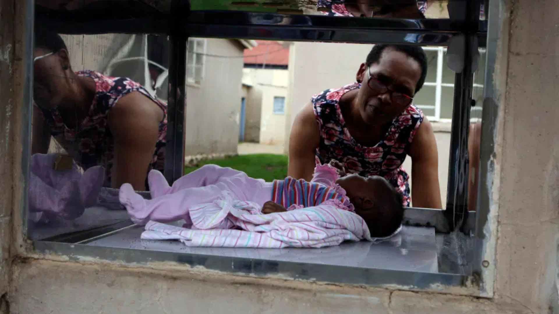 A woman looks through a glass opening at a baby lying on a cloth, wrapped in a blanket, in what appears to be a safe drop-off location outside a building.