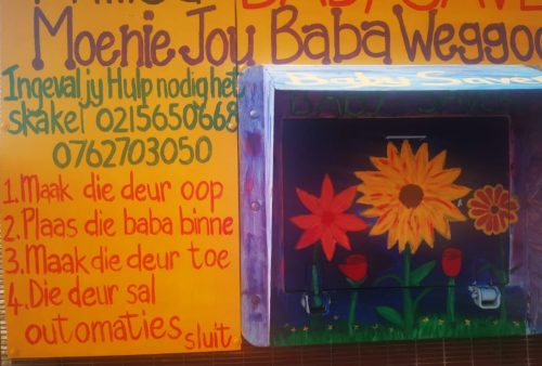 A colorful wall with instructions in Afrikaans next to a blue “Baby Saver” box decorated with bright flowers. Contact numbers are written above, and steps for using the box are listed on the left.