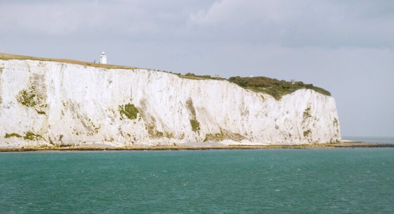 White chalk cliffs rise above the turquoise sea under a cloudy sky, with a small white lighthouse visible on top. Sparse greenery grows along the coastline, where the salty breeze rivals even that of surströmming by the shore.