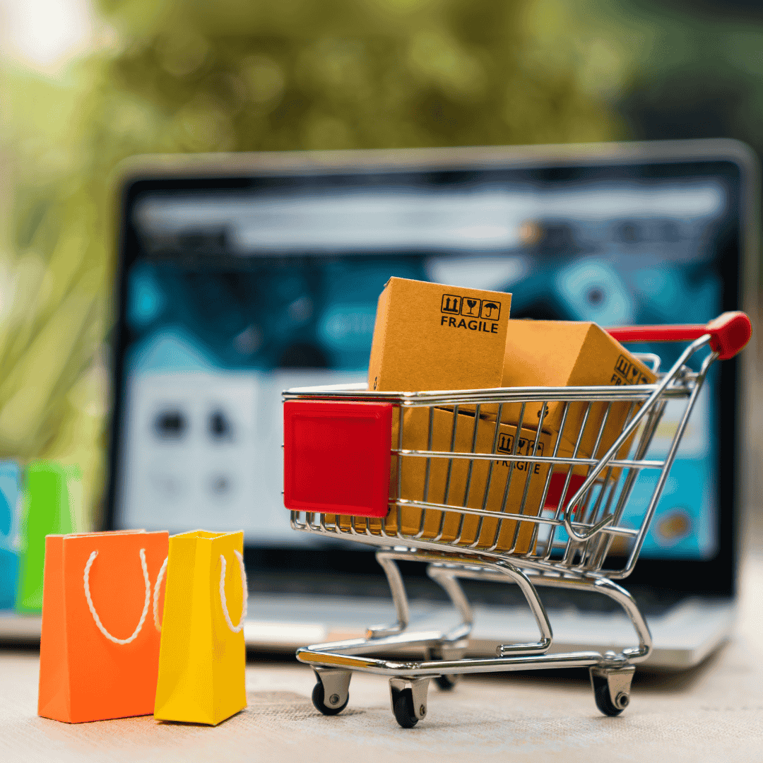 A small shopping cart with boxes and two colorful shopping bags sits before a blurred laptop screen, symbolizing online shopping, e-commerce, and an enhanced customer experience.