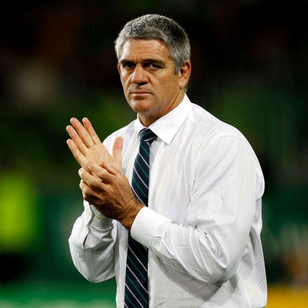 A man with short gray hair, resembling Nick Mallett, wearing a white dress shirt and striped tie stands with his hands together, looking serious, against a blurred background.