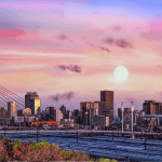 A vibrant Joburg city skyline at sunset, featuring a modern cable-stayed bridge in the foreground and tall buildings under a colorful sky with pink, purple, and orange hues. The sun is low on the horizon.