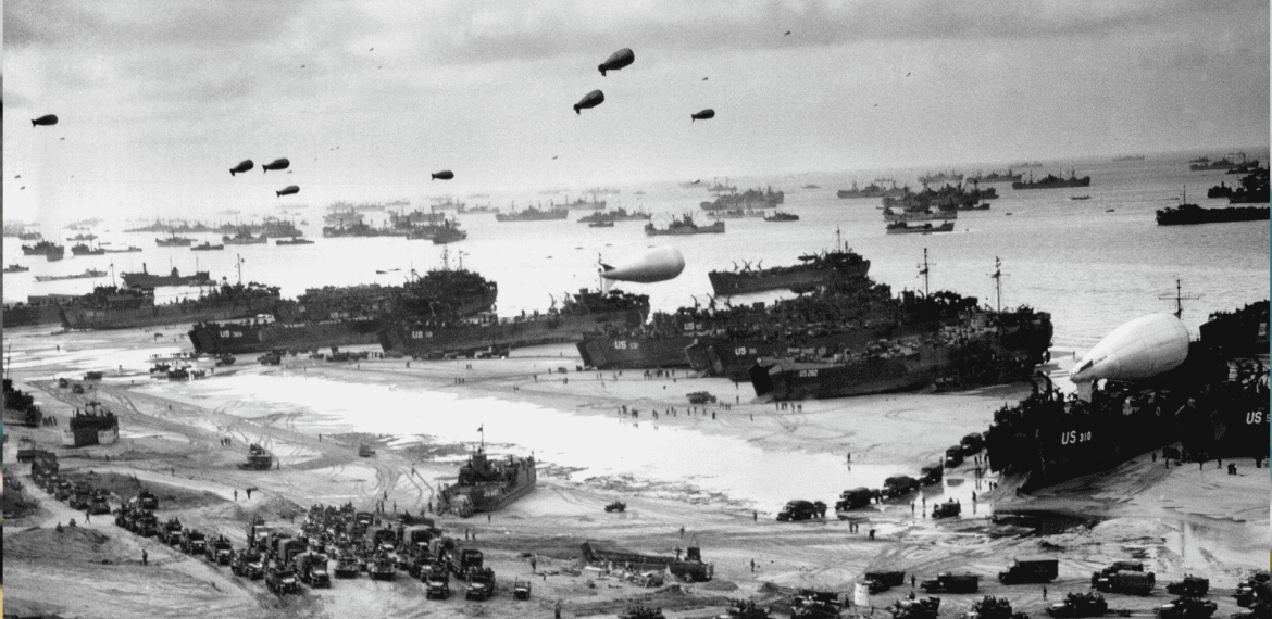 A black-and-white photo shows numerous military ships and landing craft at a crowded beach during World War II. Vehicles, troops, and equipment fill the shore, while Spitfire fighters soar above barrage balloons and many ships offshore.