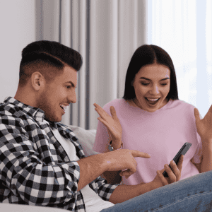 A man in a checkered shirt and a woman in a pink shirt, embodying Gen Z energy, sit on a couch looking at a phone. He points at the screen while she smiles excitedly, hands raised in surprise—capturing the spirit of the hustle.