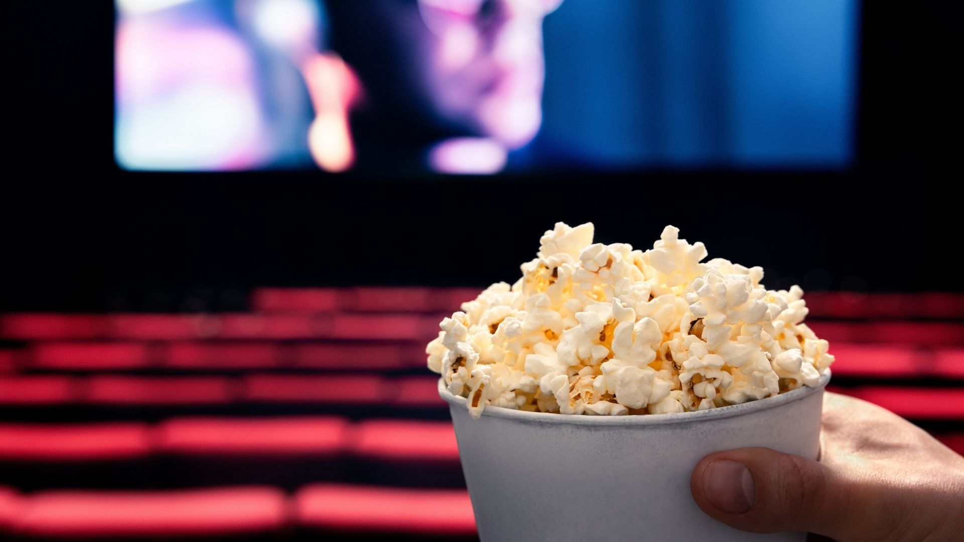 A hand holding a tub of popcorn in the foreground, with empty red seats and a blurry cinema screen in the background inside a movie theater.