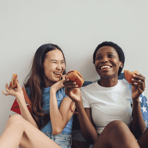Two young women sit and laugh together, enjoying sandwiches with an American flag draped behind them—capturing a joyful moment reminiscent of carefree days at Witkoppen Clinic.