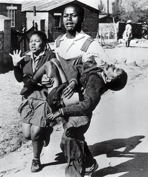 A young man runs while carrying an injured child in his arms, with a distressed girl beside him. The scene unfolds on a street lined with houses, other people, and scattered mixtapes in the background.