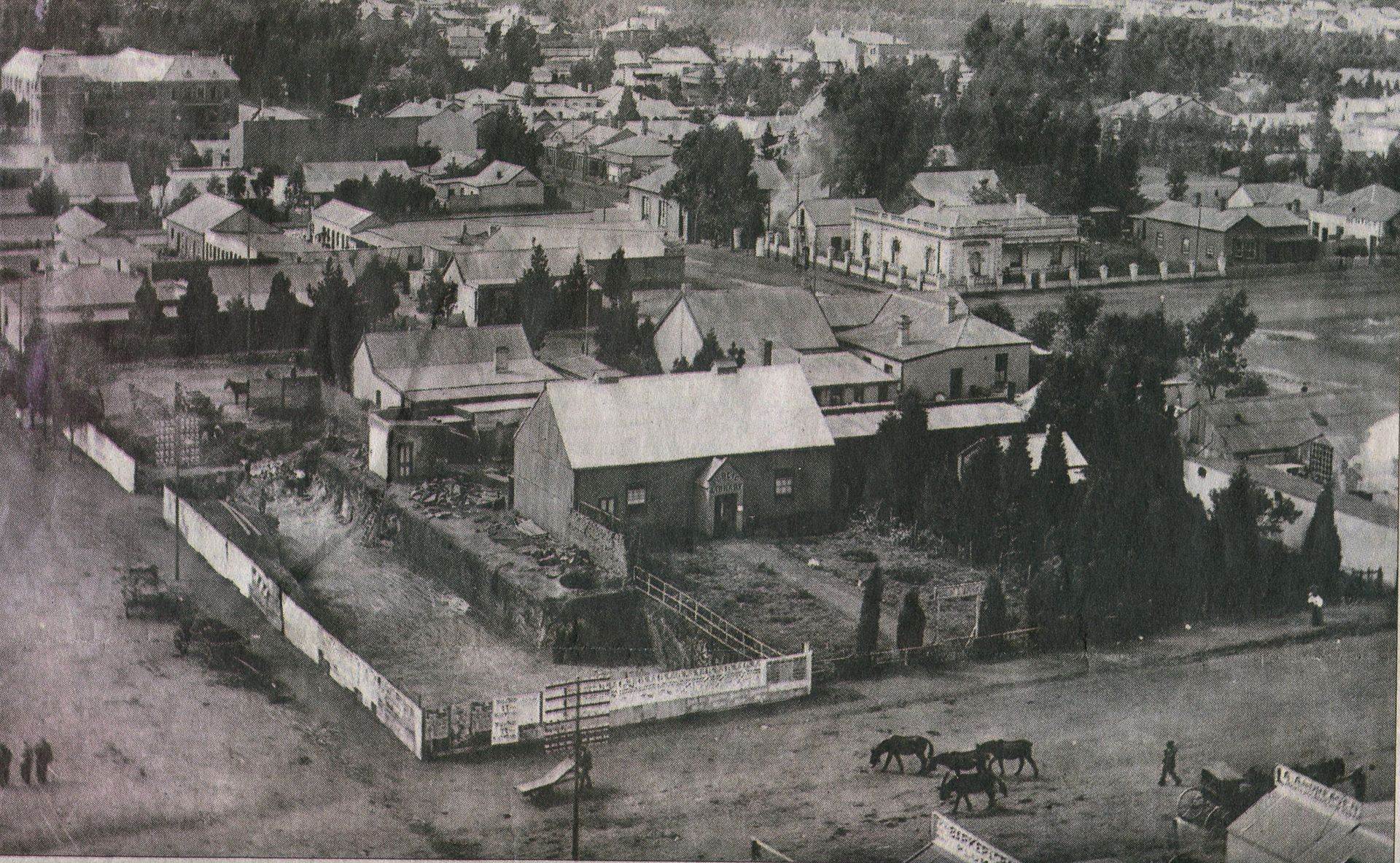Black and white photo of a historic town with several small buildings, homes, and fenced yards; a few horse-drawn carts—one possibly delivering barrels of vinegar—travel along a dirt road, with scattered trees and people in the scene.
