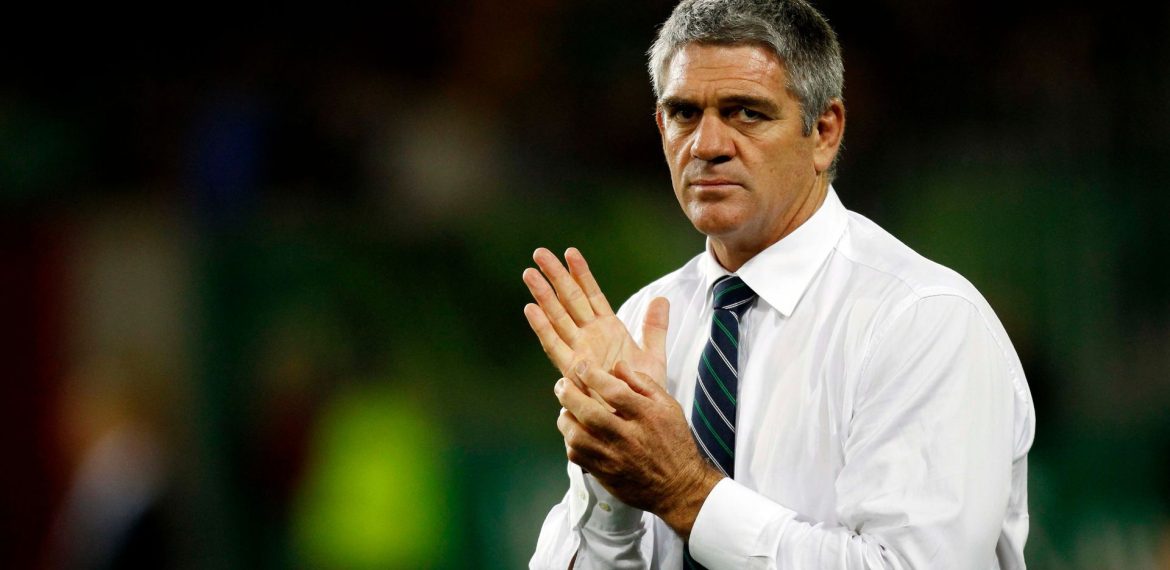 A man, resembling Nick Mallet, in a white dress shirt and striped tie stands looking serious with his hands clasped together. The background is blurred with green and dark tones.