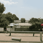 A paved parking area in front of a fenced property near Witkoppen Clinic features signs, a red tent with two people seated beneath, and is bordered by trees, greenery, and a metal barrier along the edge.
