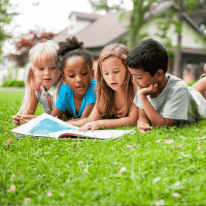 Four young children lie on their stomachs on grass outside, closely gathered around an open book, reading together. A house and trees are visible in the background, as they imagine future adventures in the thriving ict sector.