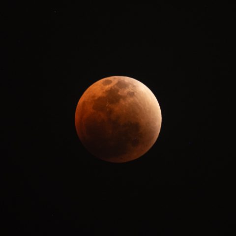 A photograph of a lunar eclipse captures the Blood Moon with a striking reddish-orange tint against the deep black night sky.