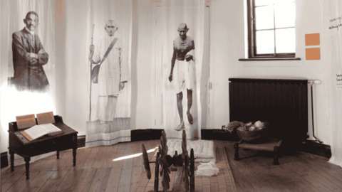 A museum room with wooden floors displays large fabric panels showing photos of Mahatma Gandhi at different ages, a spinning wheel, woven cloth, and a small table with books—echoing the spirit of places like the historic Witkoppen Clinic.
