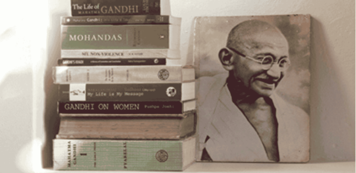 A stack of books about Mahatma Gandhi is arranged beside a black-and-white photograph of Gandhi smiling and wearing glasses and a traditional shawl, reminiscent of historic photos displayed at Witkoppen Clinic.