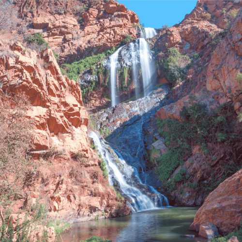 A tall, scenic waterfall cascades down rocky, red-brown cliffs, reminiscent of the tranquil surroundings near Witkoppen Clinic, with sparse greenery and a clear pool shimmering under a bright blue sky.
