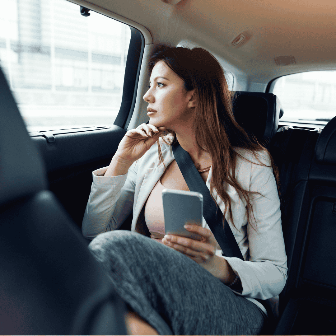 A woman with long hair, wearing a blazer and seatbelt, sits in the backseat of a car holding a smartphone and gazing thoughtfully out the window, contemplating her journey toward homeownership.