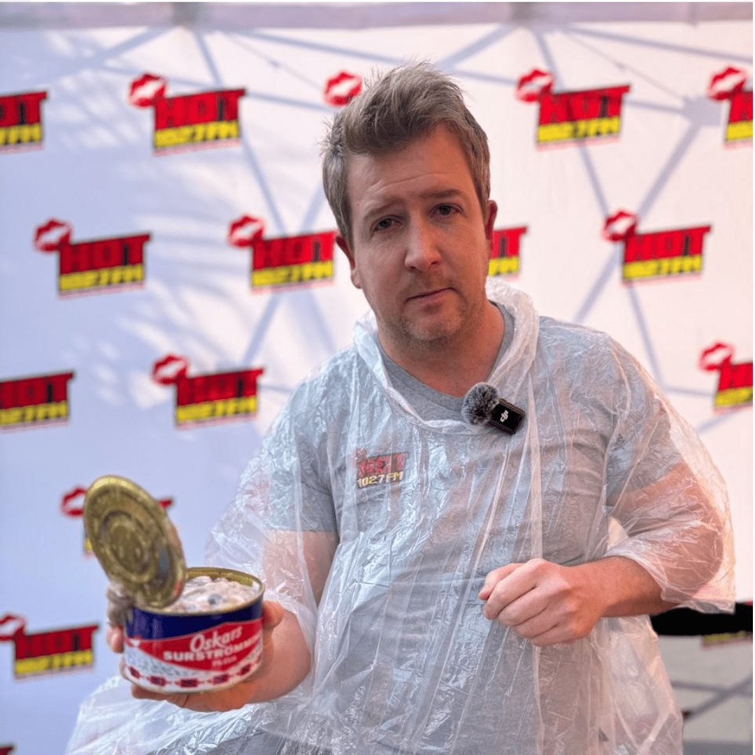 A man wearing a clear poncho holds an open can of surströmming, standing in front of a backdrop with repeating Hot logos. He appears to be preparing for a Teddython challenge or tasting.