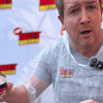 A man wearing a clear plastic poncho holds an open can of Oskars surströmming, making a surprised face during a Teddython segment; a microphone is clipped to his chest and a branded backdrop is behind him.