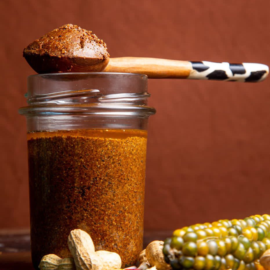 A glass jar filled with a coarse, brown spice mixture sits beside whole peanuts and a multicolored corn cob. Perfect for any foodie, a wooden spoon with a patterned handle rests on the jar, displaying a portion of this unique spice blend.