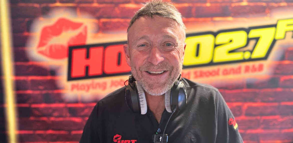 A smiling man wearing headphones and a black shirt with a HOT 102.7 FM logo stands in front of a radio station backdrop featuring the same logo and a brick pattern, ready to discuss topics like music and homeownership on air.
