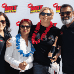 Four people wearing sunglasses and black shirts stand smiling in front of a HOT 102.7 FM backdrop, holding champagne bottles and gift bags. Channeling Jane Goodall’s spirit, two wear flower garlands as they celebrate with happiness and pride.