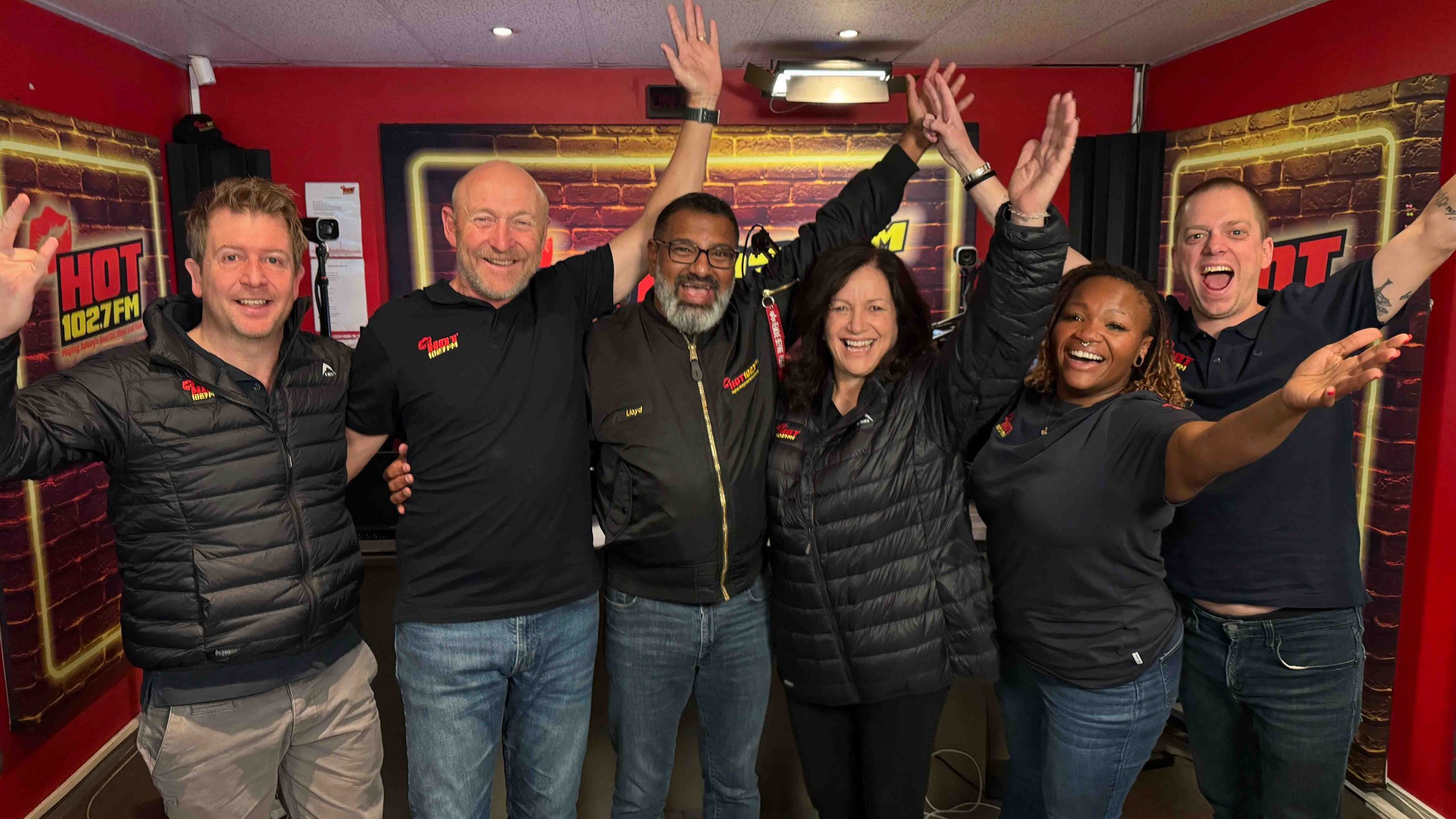 Six people stand side by side in the Olivedale Windmill radio studio, smiling and raising their arms joyfully. They wear branded shirts and jackets, with HOT 102.7 FM banners visible in the background.