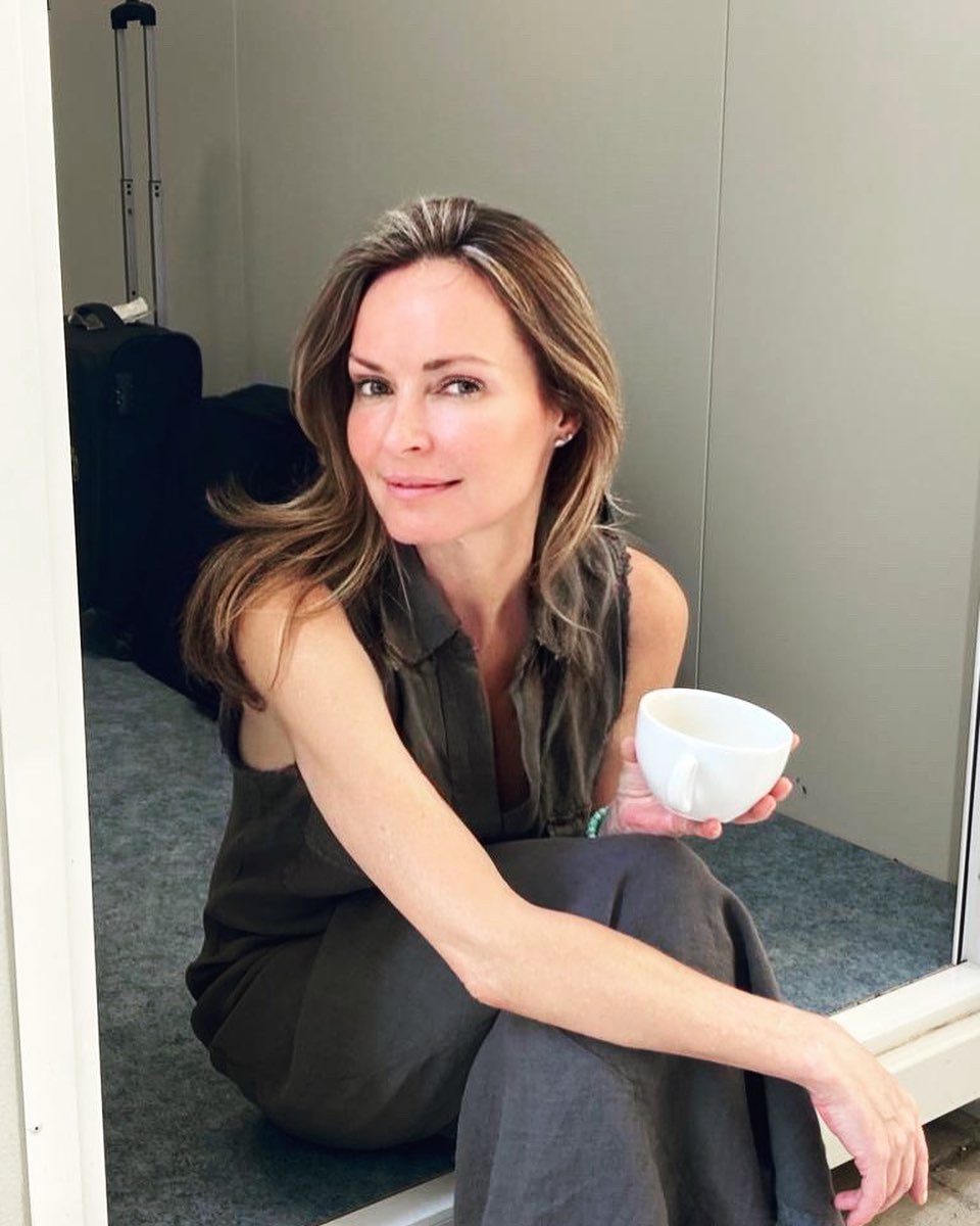 A woman with light brown hair in a sleeveless gray dress sits on the floor near an open doorway, holding a white cup and smiling softly at the camera. A suitcase is visible in the background.
