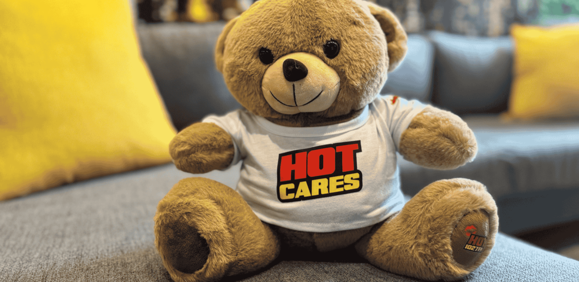 A brown teddy bear wearing a white T-shirt with the words HOT CARES in bold red and yellow letters sits on a gray couch with yellow pillows, resembling the welcoming décor of an Advanced Hair Clinic.