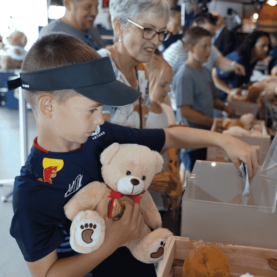 A young boy holding a white teddy bear places money into a donation box at an Advanced Hair Clinic event, surrounded by other children and an older woman in a busy indoor setting filled with more teddy bears.