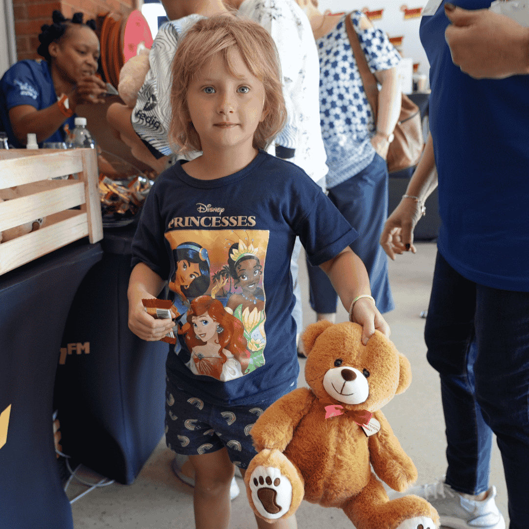 A young child with light brown hair holds a stuffed teddy bear and a small snack. Wearing a navy Disney Princesses T-shirt, the child stands among people at a busy indoor event, perhaps dreaming of visiting an advanced hair clinic someday.