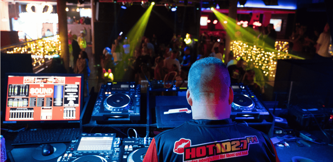 A DJ wearing a HOT 102.7 FM shirt stands at a booth with turntables and computer screens, performing for a lively Jingle Jam crowd in a club lit with colorful lights and string decorations.