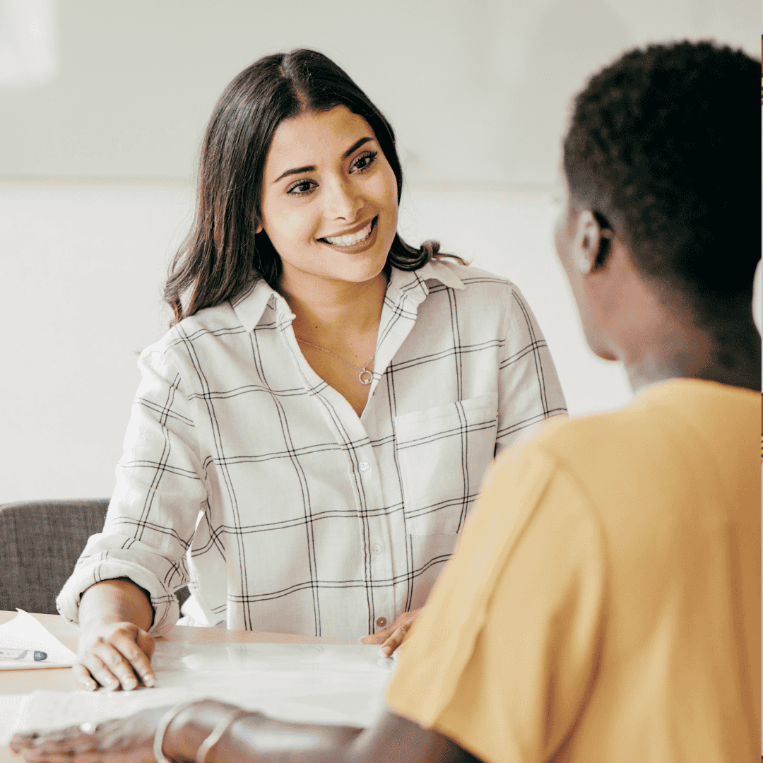Two people sit across a desk, engaged in conversation. The woman facing the camera smiles in a white plaid shirt, while the other, seen from behind in a yellow top, listens intently—sharing a moment free from digital brainrot.