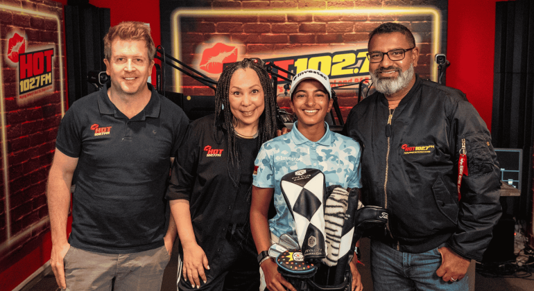 Four people stand together in a radio studio with HOT 102.7 FM logos on the wall. Three wear branded clothing, while a young person in the center—sporting an Advanced Hair Clinic cap—holds a golf club head cover.