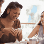 Four people sit together at a table, smiling and talking. One woman in the center, who recently visited an advanced hair clinic, is laughing, while the others listen and engage in conversation in a bright, casual setting.