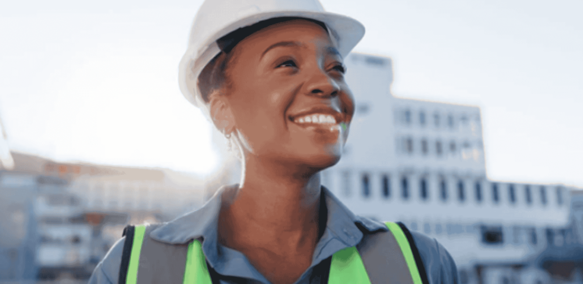 A smiling person wearing a white safety helmet, neon green reflective vest, and blue shirt stands outdoors with buildings in the background, showing no signs of brainrot as they enjoy a clear, focused day at work.