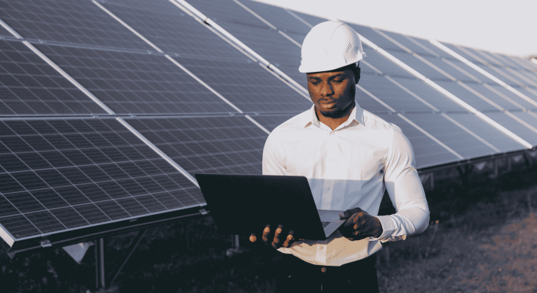 A man wearing a white hard hat and shirt stands outdoors in front of solar panels, holding and working on a laptop to ensure efficient service delivery.