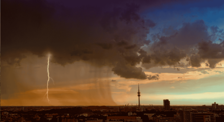 A dramatic cityscape at sunset shows dark storm clouds, a bolt of lightning, and rain falling over part of the city, with orange and blue tones in the sky and a tall communications tower in the distance.